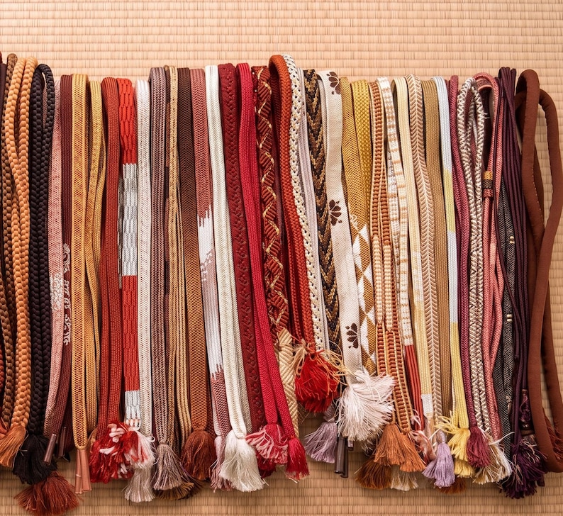 May include: A collection of colorful, decorative Japanese obi cords. The cords vary in color, texture, and design, including braided, patterned, and tasseled styles. Colors include red, brown, gold, and white. The cords are arranged vertically against a neutral background.