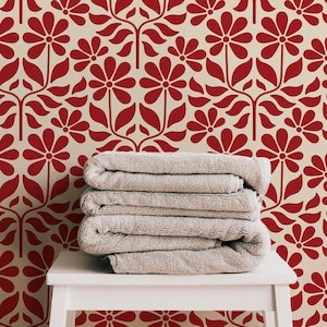 Può includere: Un mucchio di quattro asciugamani beige piegati ordinatamente su un tavolo bianco davanti a una carta da parati floreale rossa e bianca.