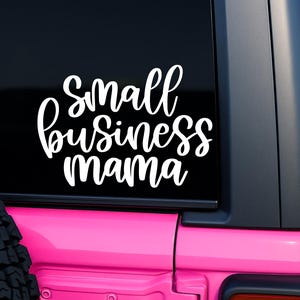以下が含まれることがあります： ピンク色の車のリアウィンドウに、筆記体フォントで「Small Business Mama」と書かれた白いビニールステッカーが貼られています。ステッカーは黒い窓の中央に配置され、車のピンク色のボディが下に表示されています。