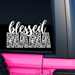 May include: White vinyl decal with the words "blessed MAMA" in a script font and leopard print. The decal is displayed on a black window of a pink vehicle.