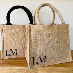 May include: Two jute tote bags, one with a black handle and one with a natural-coloured handle. Each bag features the initials "LM" printed in black. The bags are made from a woven, natural material.