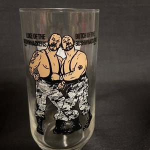 May include: A clear glass with a black and white illustration of two wrestlers, Luke and Butch of the Bushwhackers, wearing camouflage pants and boots. The text "Luke of the Bushwhackers" and "Butch of the Bushwhackers" is printed on the glass.