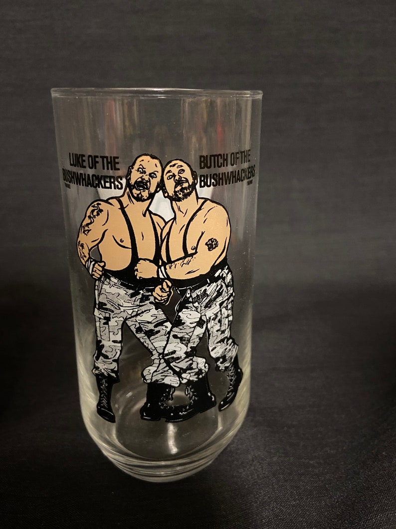May include: A clear glass with a black and white illustration of two wrestlers, Luke and Butch of the Bushwhackers, wearing camouflage pants and boots. The text "Luke of the Bushwhackers" and "Butch of the Bushwhackers" is printed on the glass.