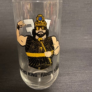 May include: A clear glass with a black and gold image of a wrestler wearing a crown and sunglasses. The text "Macho King" Randy Savage is printed on the glass.