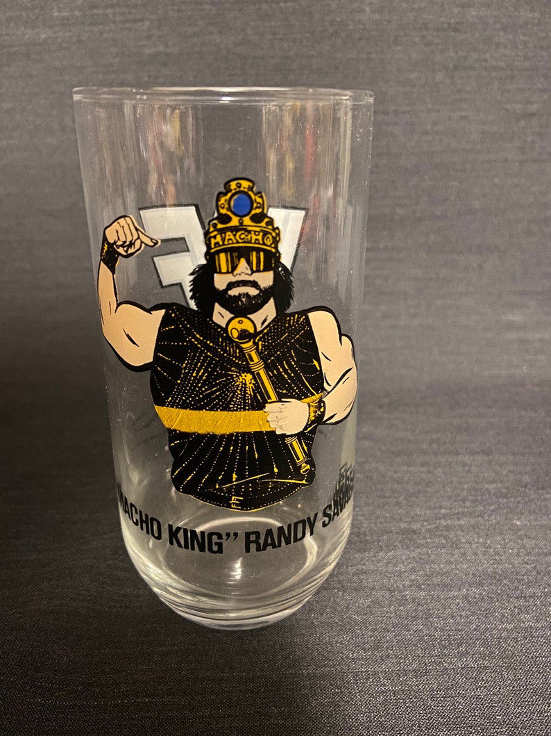 May include: A clear glass with a black and gold image of a wrestler wearing a crown and sunglasses. The text "Macho King" Randy Savage is printed on the glass.