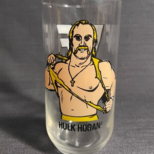May include: A clear glass with a cartoon image of Hulk Hogan wearing a yellow wrestling outfit and a gold chain with a cross pendant. The glass has the text "HULK HOGAN" printed on the bottom.