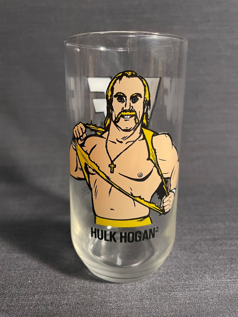 May include: A clear glass with a cartoon image of Hulk Hogan wearing a yellow wrestling outfit and a gold chain with a cross pendant. The glass has the text "HULK HOGAN" printed on the bottom.