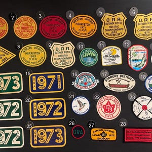 May include: A collection of 28 colourful patches with various designs and text. The patches are numbered 1 through 28 and feature themes such as "O.R.A. Outdoor Pistol Championships", "Fall Outdoor", "Western Ontario Harrison", "Sudbury District Voyageurs", "Silver Expert Shield Rifle", "National Matches Canada 1972", and "East Canada Outdoor Handgun Championships".