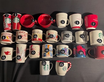 May include: A collection of 23 ceramic mugs with various designs and colours. The mugs are numbered 1 through 23. Some mugs feature city names such as "California", "Manila", and "Washington".
