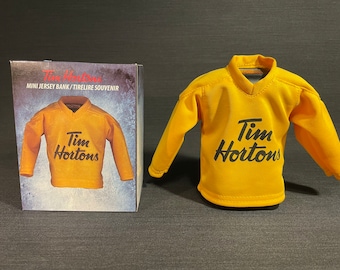 May include: A yellow Tim Hortons mini jersey bank. The jersey has black lettering that says "Tim Hortons".