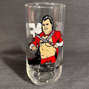 May include: A clear glass with a red and black illustration of a wrestler holding scissors. The text "BRUTUS "THE BARBER" BEEF" is printed on the glass.