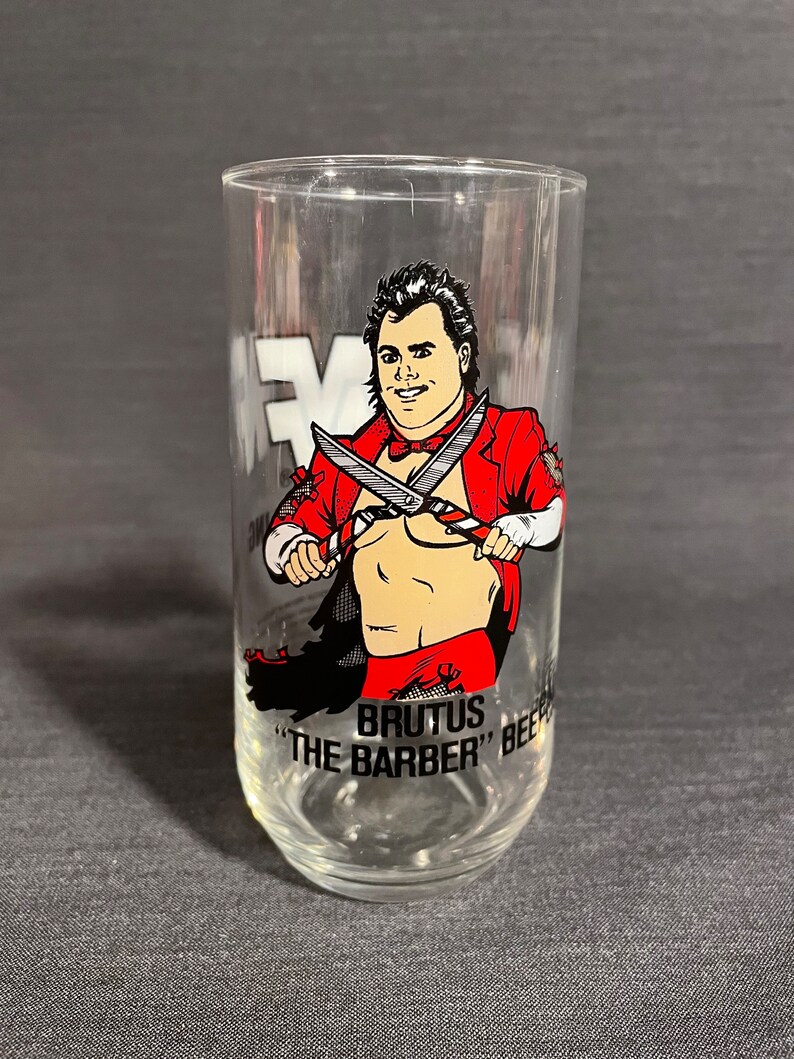 May include: A clear glass with a red and black illustration of a wrestler holding scissors. The text "BRUTUS "THE BARBER" BEEF" is printed on the glass.