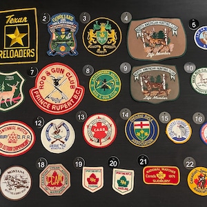 May include: A collection of 22 embroidered patches, mostly featuring hunting and fishing themes. The patches are arranged in rows on a black background. Some of the patches feature text, such as "Texan Reloaders", "North American Hunting Club", and "Rod & Gun Club".