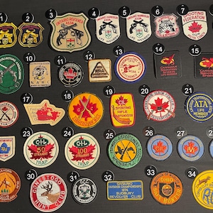 May include: A collection of 34 colourful embroidered patches featuring various shooting and hunting clubs and organisations. The patches are arranged in rows on a black background. Some of the patches feature maple leaves, rifles, and other hunting-related imagery.