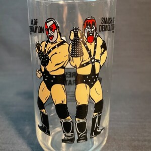 May include: A clear glass with a black and white image of two wrestlers. The wrestlers are wearing black wrestling gear and have red and black face paint. The text "Ax of Demolition" and "Smash of Demolition" is printed on the glass.