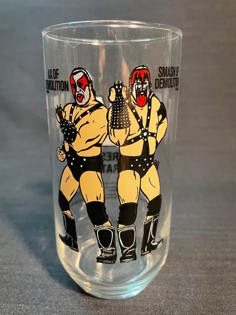May include: A clear glass with a black and white image of two wrestlers. The wrestlers are wearing black wrestling gear and have red and black face paint. The text "Ax of Demolition" and "Smash of Demolition" is printed on the glass.
