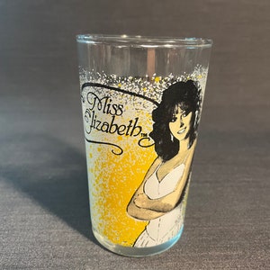May include: A clear glass with a yellow and white splatter design. The glass features a black and white illustration of a woman with long black hair. The text "Miss Elizabeth" is printed on the glass.
