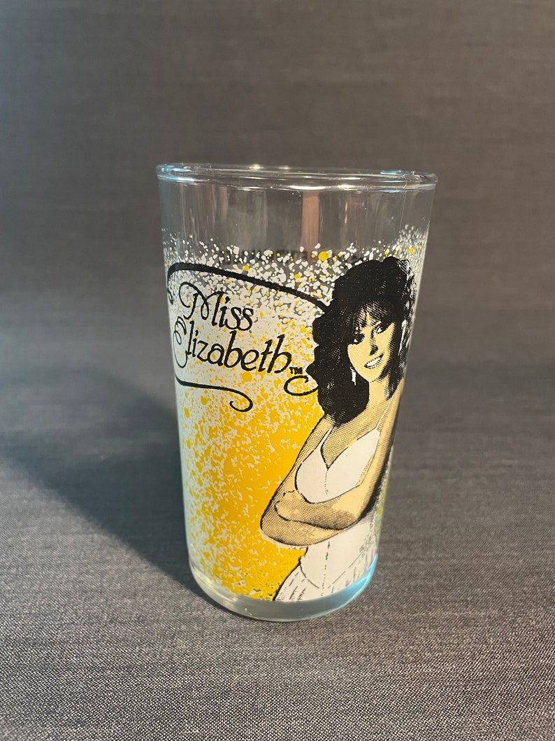 May include: A clear glass with a yellow and white splatter design. The glass features a black and white illustration of a woman with long black hair. The text "Miss Elizabeth" is printed on the glass.