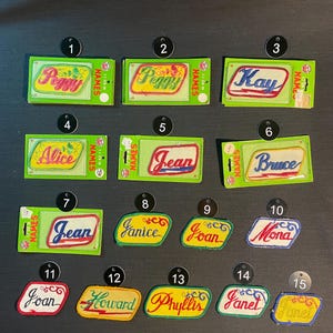 May include: Fifteen colorful iron-on name patches with various names including Peggy, Kay, Alice, Jean, Bruce, Joan, Janice, Mona, Howard, Phyllis, and Janet. The patches are on green and white cards with the word "NAMES" printed on them.