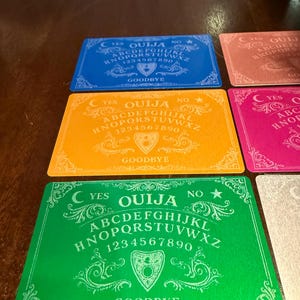May include: A collection of colorful Ouija board planchette cards in blue, orange, green, and pink. Each card features the alphabet, numbers, and the words "Yes", "No", and "Goodbye". The cards have decorative scrollwork.