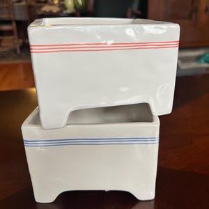 May include: Two white ceramic square planters. The top planter has two red stripes, and the bottom planter has two blue stripes. The planters have a rectangular base with a small footed design.