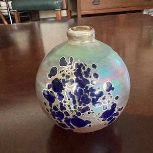 May include: A spherical, iridescent glass vase with a short neck. The vase is decorated with a pattern of dark blue, irregular shapes. The glass has a subtle, colourful sheen, reflecting light.
