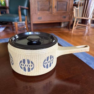 May include: A tan ceramic cooking pot with a black lid and handle. The pot features a repeating blue and white design around the exterior. The pot is sitting on a dark brown wooden table.