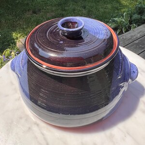Op de afbeelding: Een grote, donkerblauwe en zwarte keramische pot met een deksel. De pot heeft een rode rand en een blauwe glazuur. Het deksel heeft een kleine knop bovenop.