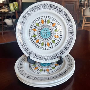 May include: A stack of white ceramic plates with a colorful geometric design. Each plate features a central turquoise and orange pattern surrounded by black and white circular and diamond shapes. The plates are stacked on a black plate stand.
