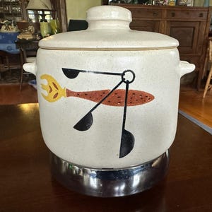 Peut inclure: Un bocal à biscuits en céramique vintage avec un couvercle, présentant une finition crème mouchetée. Le bocal est orné d'un motif rétro d'une fourchette jaune, d'un ovale marron et de formes noires en forme de cuillère. Il repose sur une base chromée.