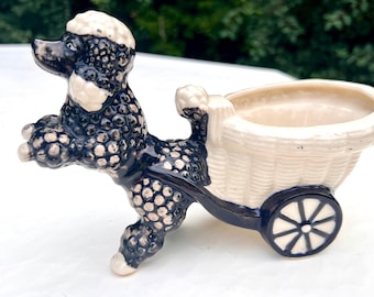 May include: A black and white ceramic poodle figurine pulling a white planter with a blue rim and a single blue wheel.