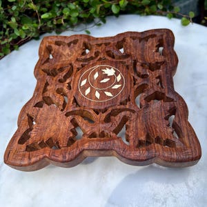 Puede incluir: Una caja de madera cuadrada, tallada intrincadamente, con un acabado marrón oscuro. El diseño presenta patrones calados de hojas y motivos florales. El centro tiene una incrustación circular con un diseño de hoja.