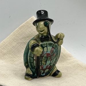 May include: A ceramic figurine of a turtle wearing a top hat, bow tie, and monocle. The turtle is holding a cane and has a green, blue, and red shell.