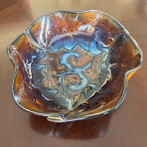 Puede incluir: Un cuenco de cerámica decorativo con una forma orgánica única. El cuenco presenta un esmalte brillante con una mezcla de tonos marrones, naranjas y azules. El borde tiene un borde oscuro. El interior tiene un patrón arremolinado.