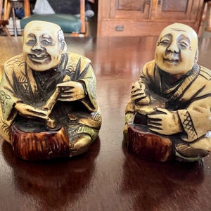 May include: Two seated figurines with smiling faces. The figures are dressed in patterned robes with tan, green, and brown tones. Each figure sits on a brown, tree-stump-shaped base. One figure holds a flute, and the other holds a small object.