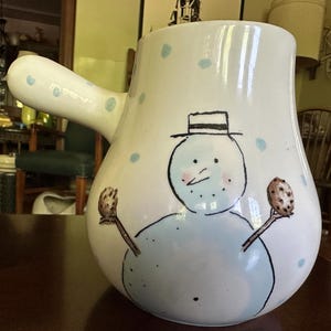 Puede incluir: Una jarra de cerámica blanca con un asa lateral, que presenta una ilustración pintada a mano de un muñeco de nieve con un sombrero de líneas negras. El muñeco sostiene una galleta marrón en cada mano, rodeado de lunares azul claro.