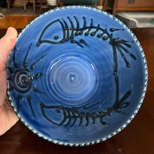 Puede incluir: Un cuenco de cerámica azul con un diseño de esqueleto de pez. El cuenco tiene una superficie texturizada y un borde azul oscuro. El diseño del esqueleto de pez es negro. El cuenco está sostenido por una mano.