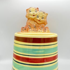Op de afbeelding: Een vintage keramische koekjespot met een gestreept ontwerp in rood, geel en blauw. Het deksel heeft twee schattige tijgerwelpjes die samen zitten.