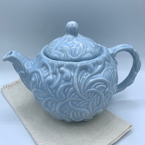 May include: A light blue ceramic teapot with an intricate floral design. The teapot has a lid and a spout. It is sitting on a white linen napkin.