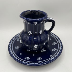 Könnte beinhalten: Ein kleiner blauer und weißer Keramik Krug mit Henkel und einer passenden Untertasse. Krug und Untertasse sind mit einem traditionellen deutschen Volkskunst-Muster aus weißen Blumen auf dunkelblauem Hintergrund verziert.