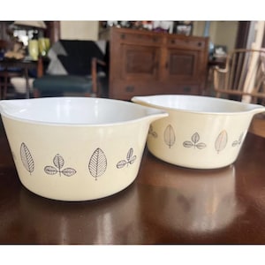 May include: Two vintage Pyrex mixing bowls. The bowls are a pale yellow with a white interior. Each bowl features a black leaf pattern around the exterior. The bowls are sitting on a dark brown table.