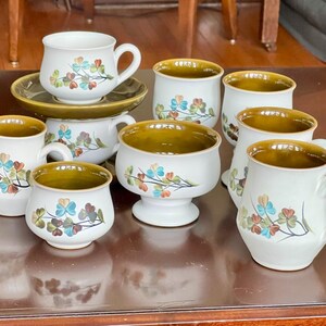 May include: A collection of white ceramic dishware with a floral design in shades of blue, orange, and green. The set includes mugs, bowls, and a creamer, all with a brown interior and rim.