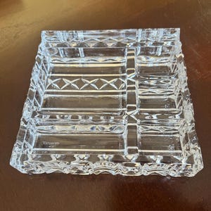 May include: A clear, rectangular Waterford crystal dish with a geometric design. The dish features a grid pattern with diamond-shaped accents and the brand name etched on the front. The edges are textured, adding to its elegant appearance.