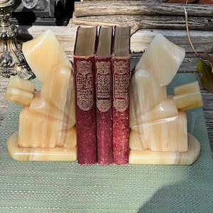 Peut inclure: Trois livres anciens avec des couvertures bordeaux, maintenus entre deux serre-livres en onyx sculpté. Les serre-livres sont de couleur ambre clair et présentent un design sculpté unique. Les livres ont des lettres dorées et sont intitulés "The Tragedies of William Shakespeare" et "The Poems of Alfred Tennyson."