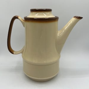 Puede incluir: Una cafetera de cerámica de color crema con un borde y un asa marrones. La cafetera tiene un diseño texturizado que se asemeja al bambú.