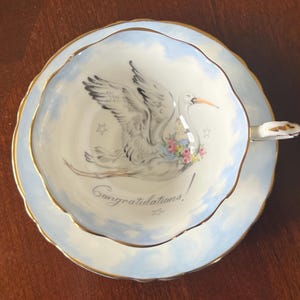 May include: A white teacup and saucer with a blue and white cloud design and gold trim. The teacup features a painted image of a stork carrying a baby in a basket with a floral wreath. The text "Congratulations!" is written on the teacup.
