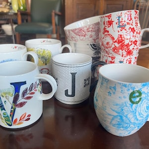 Op de afbeelding: Een verzameling keramische mokken met diverse ontwerpen. Eén mok heeft een grote bloemen "V", een andere heeft een "J" op een zwart-wit raster, en andere tonen bloemen- en patroonmotieven. De mokken zijn wit met kleurrijke accenten.