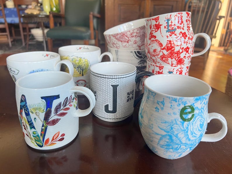 K&ouml;nnte beinhalten: Eine Sammlung von Keramikbechern mit verschiedenen Designs. Ein Becher zeigt ein gro&szlig;es florales "V", ein anderer ein "J" auf einem schwarz-wei&szlig;en Gitter, und andere zeigen florale und gemusterte Motive. Die Becher sind wei&szlig; mit farbigen Akzenten.