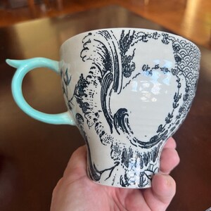 K&ouml;nnte beinhalten: Eine wei&szlig;e Keramiktasse mit einem schwarzen Wellen- und Blumendesign. Die Tasse hat einen hellblauen Griff und ein einzigartiges, geschwungenes Design. Die Tasse wird in einer Hand gehalten.