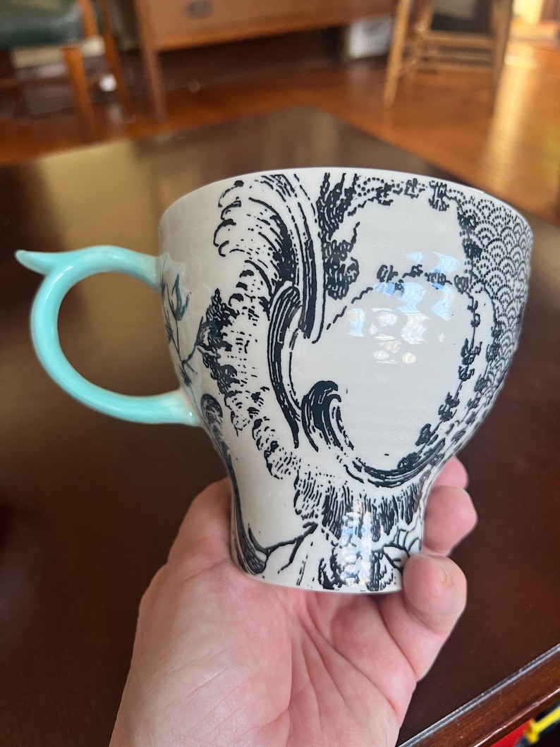 K&ouml;nnte beinhalten: Eine wei&szlig;e Keramiktasse mit einem schwarzen Wellen- und Blumendesign. Die Tasse hat einen hellblauen Griff und ein einzigartiges, geschwungenes Design. Die Tasse wird in einer Hand gehalten.